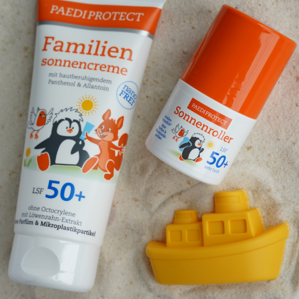 Die Familiensonnencreme befindet sich auf der linken Seite des Bildes. Der Sonnenroller befindet sich auf der oberen rechten Seite und das Schiff-Sandförmchen befindet sich auf der unteren rechten Seite des Bildes. Die Produkte liegen auf dem Sand.
