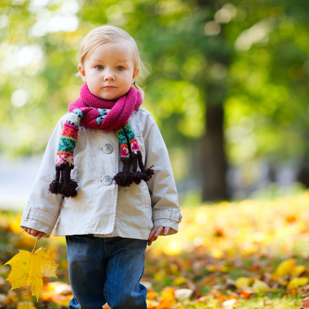 kleines Mädchen, das mit Jacke und Schal durch das Herbstlaub läuft 